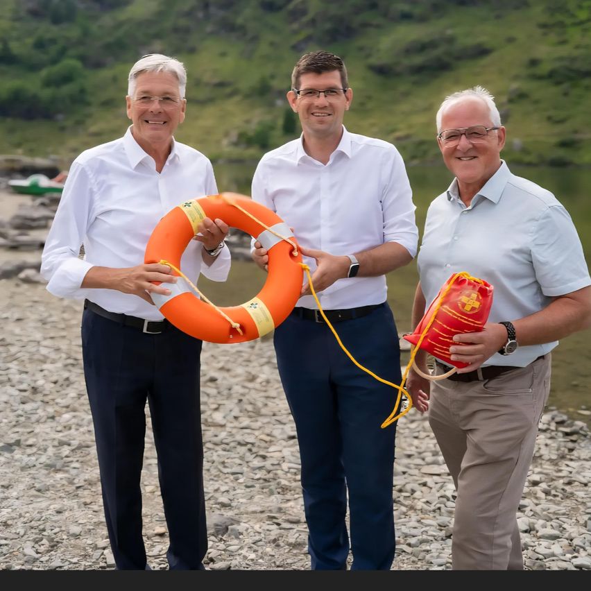 Drei Männer stehen am felsigen Ufer. Einer hält eine Rettungsboje, ein anderer eine Rettungsbojentasche. Im Hintergrund ist ein Fluss und grüne Hügel zu sehen.
