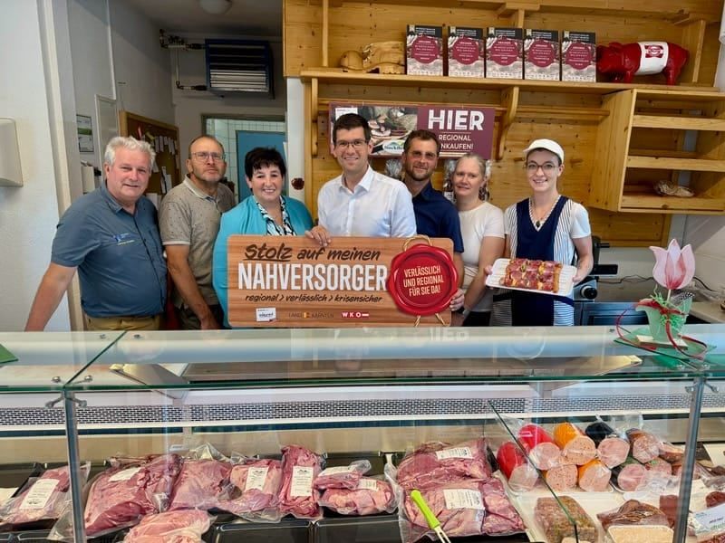 Eine Gruppe von Menschen steht hinter einer Fleischtheke. Sie halten ein Schild mit der Aufschrift 'Stolz auf meinen Metzger'. Im Vordergrund sind verschiedene Fleischsorten ausgestellt.