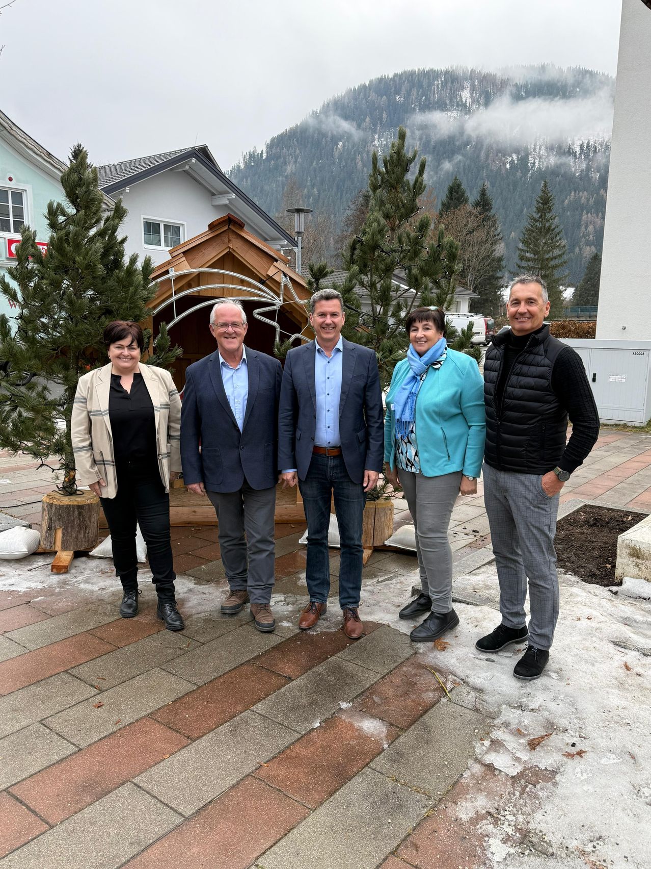Fünf Menschen stehen lächelnd auf einem gepflasterten Weg vor einem verschneiten Berg. Zwei Frauen und drei Männer, alle schick gekleidet. Hinter ihnen stehen Weihnachtsbäume und Häuser.