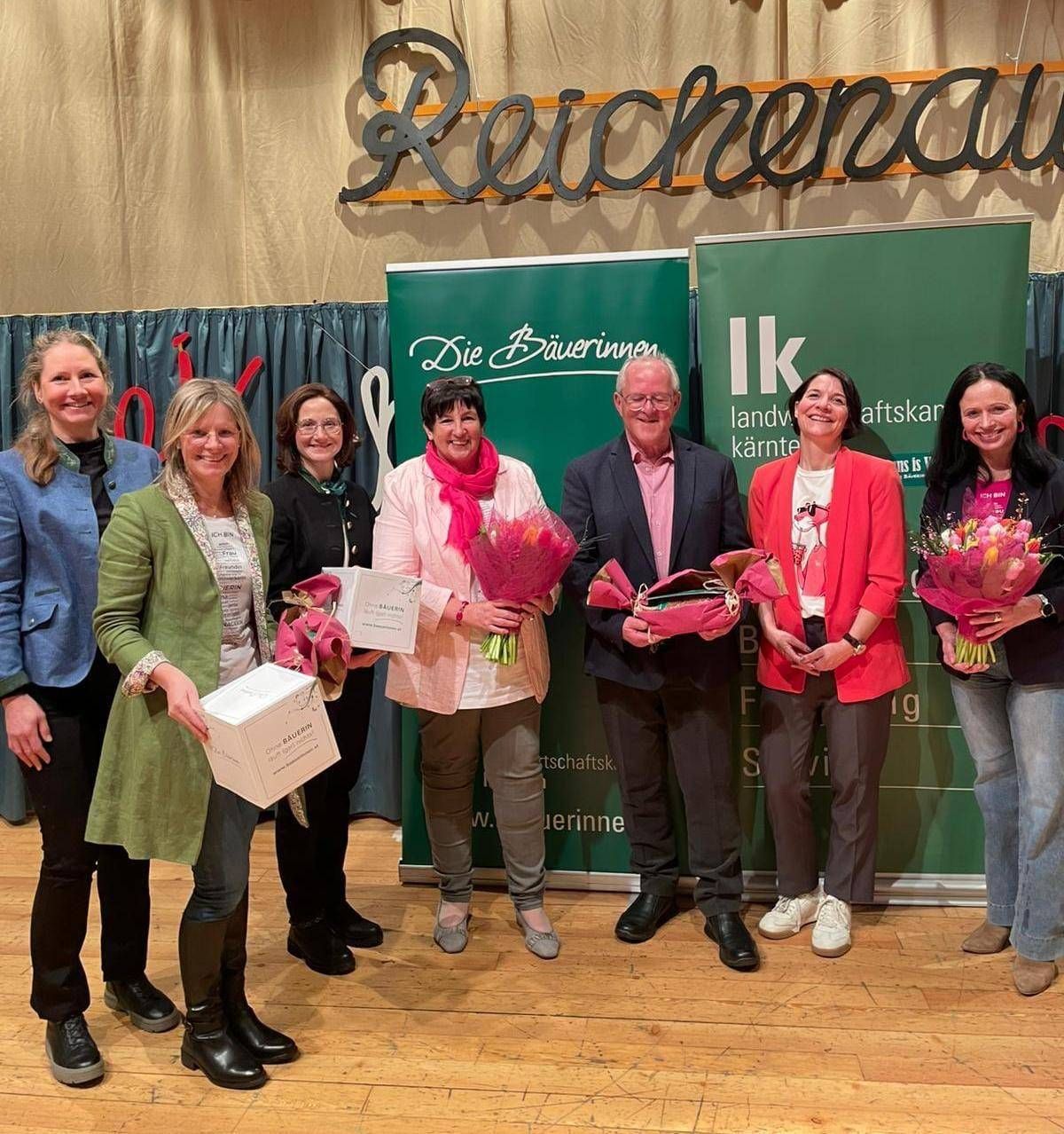 Eine Gruppe von Menschen steht vor einem Banner mit einer Frau, die Blumen hält und lächelt. Sie scheinen Auszeichnungen zu erhalten.