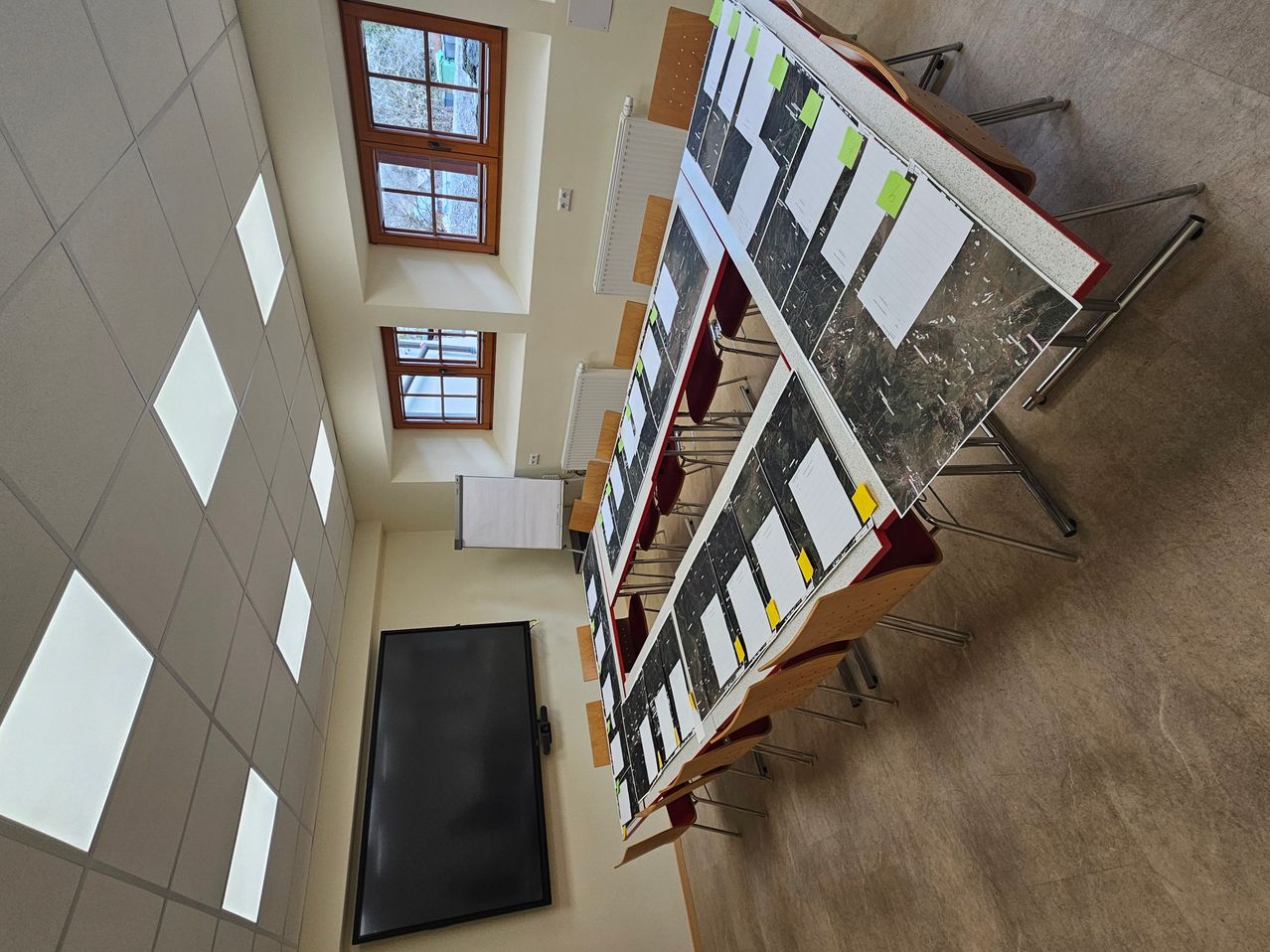 Eine Vogelperspektive eines Klassenzimmers mit einem langen Tisch, Stühlen und Papieren. Fenster und eine Tafel sind an der Wand.