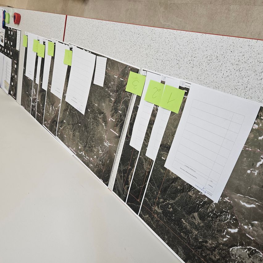 Eine Reihe von Karten ist an einer weißen Tafel befestigt, mit grünen Haftnotizen versehen. Die Karten sind schwarz-weiß und zeigen verschiedene geografische Merkmale.