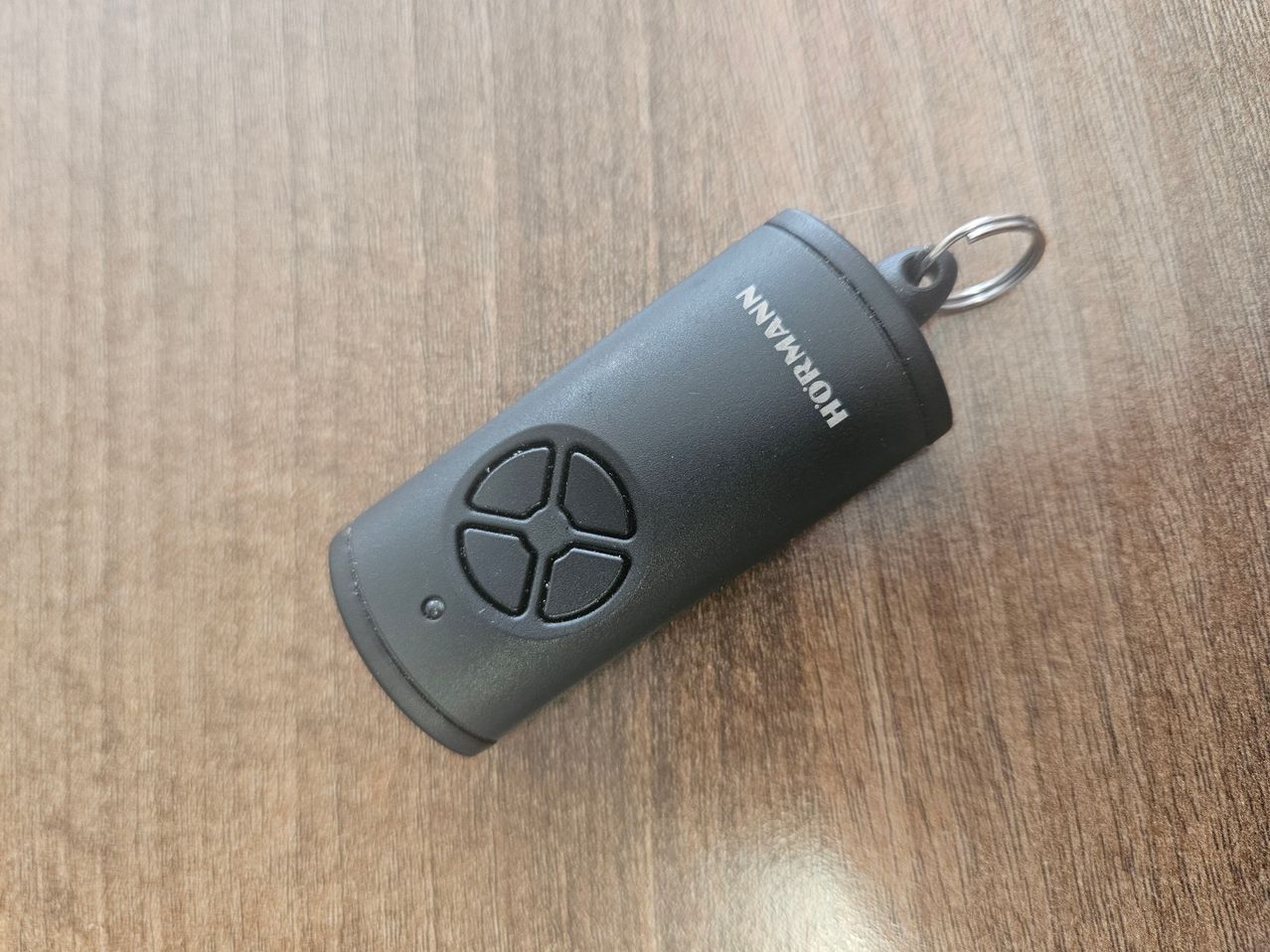 Ein schwarzer, zylindrischer Fernbedienung mit einem silbernen Schlüsselring liegt auf einer Holzoberfläche. Die Fernbedienung hat eine kreisförmige Taste in der Mitte und den Schriftzug 'HONEYWELL' darauf.