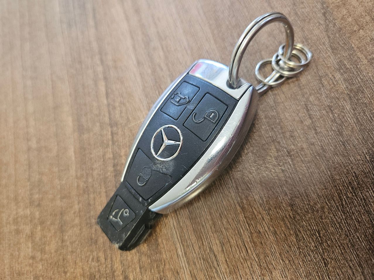 Ein Mercedes-Benz Autoschlüssel mit silbernem Metallgehäuse, schwarzen Tasten und einem silbernen Schlüsselring auf einer Holzoberfläche.