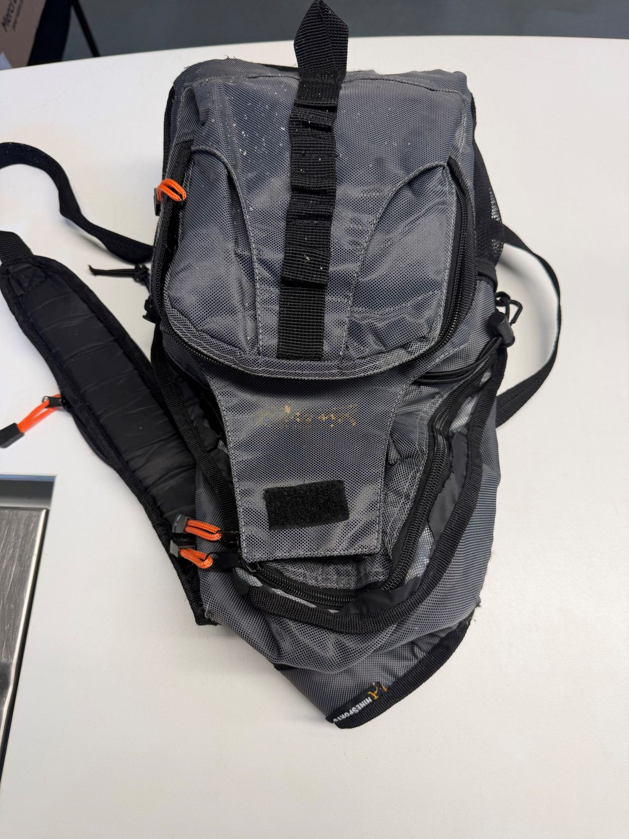 A close-up view of a gray backpack with black and orange straps and zippers, resting on a white surface.