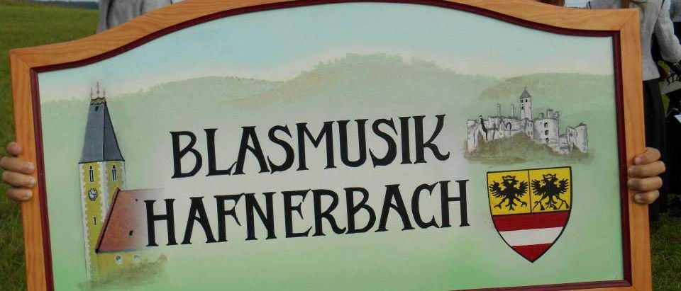 Eine Frau hält ein Holzschild mit dem Wappen und Namen der Stadt 'Blasmusik Hafnerbach', das eine Kirche und eine Burg zeigt.