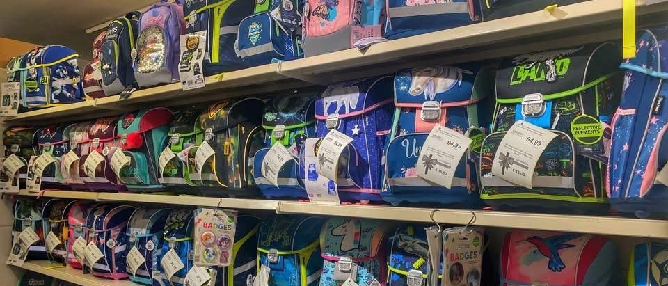 Laden gefüllt mit bunten Rucksäcken, meist mit Etiketten und Preisschildern. Einige haben das Wort Abzeichen darauf. Die Regale sind ordentlich organisiert und mit verschiedenen Rucksäcken gefüllt.