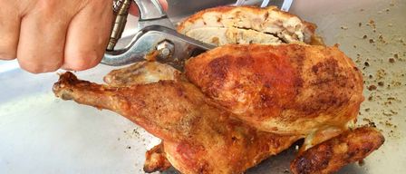 Eine Person benutzt eine Küchenschere, um ein gebratenes Huhn auf einem Metalltablett zu schneiden. Der Text darüber lautet 'Sonntag ist wieder Steirerhuhn-Grill'.