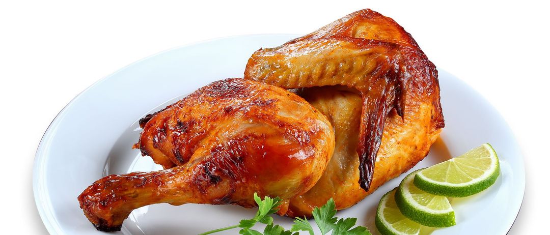 Ein weißer Teller mit einem gebratenen Huhn, knuspriger Haut und mit Petersilie und Limettenscheiben garniert. Das Huhn ist in halben Positionen serviert und zeigt sein saftiges Inneres.