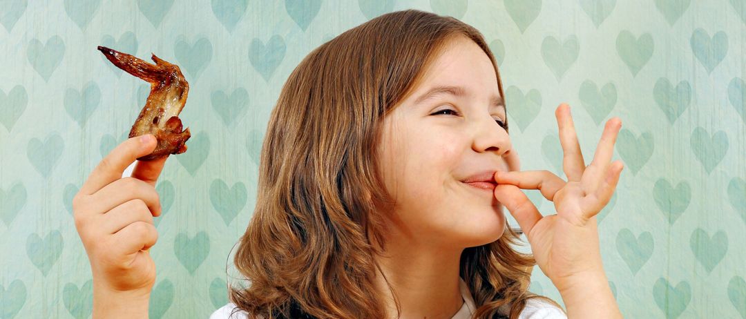 Ein junges Mädchen lächelt, während es einen Hähnchenflügel hält und für ein Foto posiert, mit einer Wand aus blauen Herzmustern im Hintergrund.