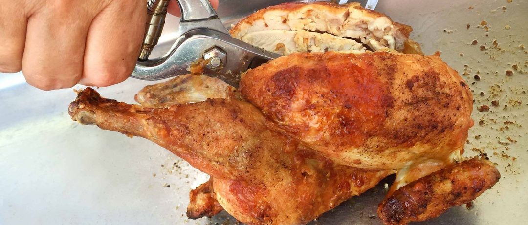 Eine Hand benutzt einen metallenen Nussknacker, um ein gebratenes Huhn zu schneiden, das auf einem Metalltablett liegt. Das Huhn hat eine goldbraune Haut und knusprige Textur. Ein Gabel ist neben dem Huhn. Das Tablett liegt auf einer weißen Oberfläche.