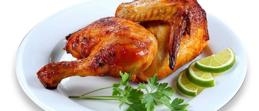 Ein weißer Teller mit einem Hähnchenschenkel und -flügel, garniert mit Petersilie und Limettenscheiben. Das Hähnchen ist goldbraun, was auf eine gute Zubereitung hinweist.