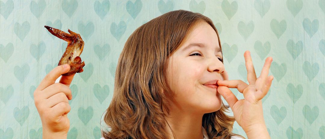 Ein Mädchen hält einen frittierten Hühnerflügel und macht mit ihren Fingern ein Friedenszeichen, während sie lächelt. Sie steht vor einer Wand mit einem Muster aus blauen Herzen.