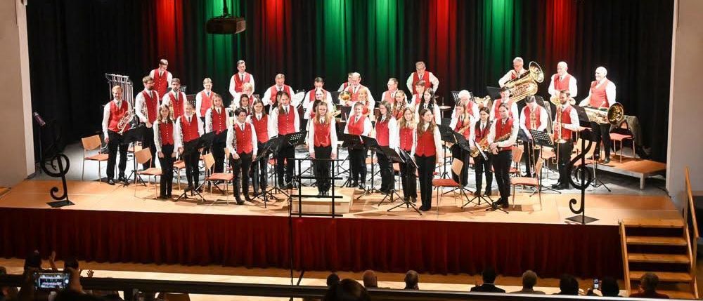 Ein Blasorchester spielt auf einer Bühne mit roten und grünen Vorhängen. Die Musiker stehen in Reihen, und einige spielen Trompeten. Das Publikum sitzt in den Stühlen vor der Bühne.