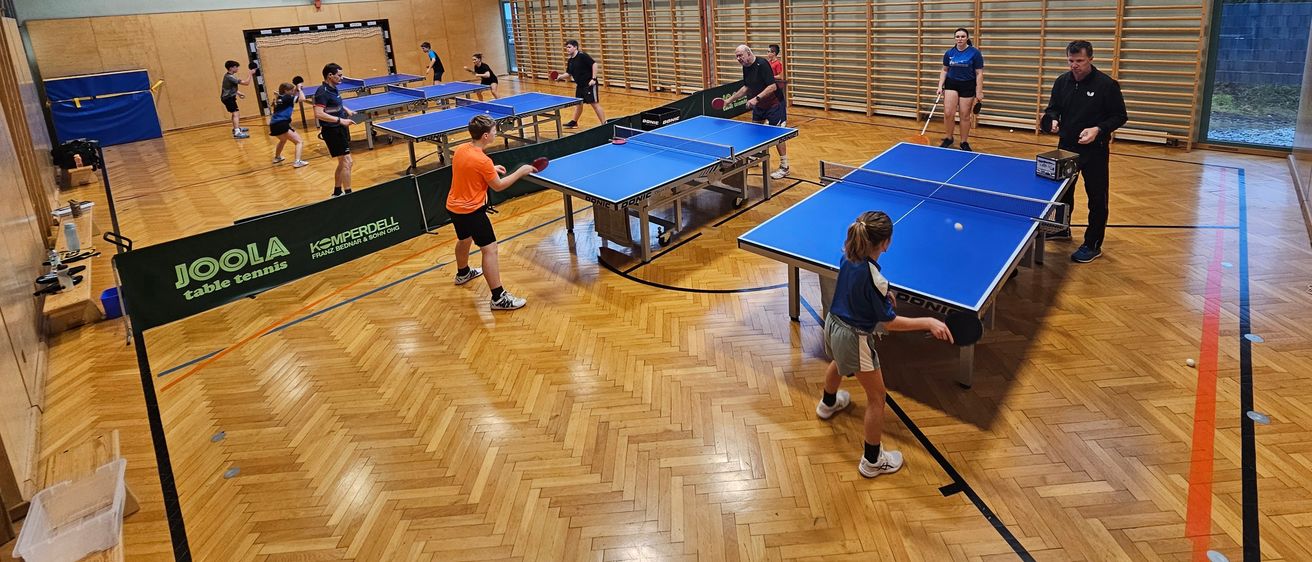 Überdachter Tischtennisraum mit mehreren Spielern, die an einem Spiel teilnehmen. Der Platz hat blaue Tische und eine Anzeigetafel ist sichtbar. Die Spieler tragen Freizeitkleidung.