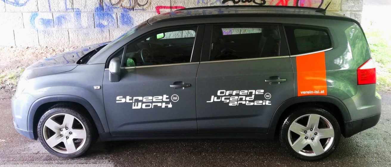 Ein graues Auto mit orangefarbenen Akzenten ist unter einer Betonbrücke mit Graffiti geparkt. Das Auto hat 'Street Work' und 'Offene Jugendarbeit' Aufkleber.