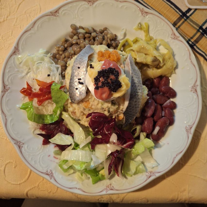 Ein Teller mit einem Fischkopf, verziert mit Kaviar, umgeben von Bohnen, Rotkohl, Salat und einer Tomate auf einer gemusterten Tischdecke.