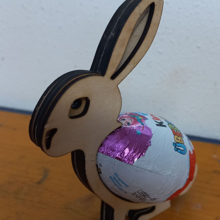 Eine hölzerne Hasenfigur hält ein purpurfarben umwickeltes Kinderüberraschungsei. Das Ei hat weiße und rote Schwünge. Der Hase steht auf einer Holzoberfläche.