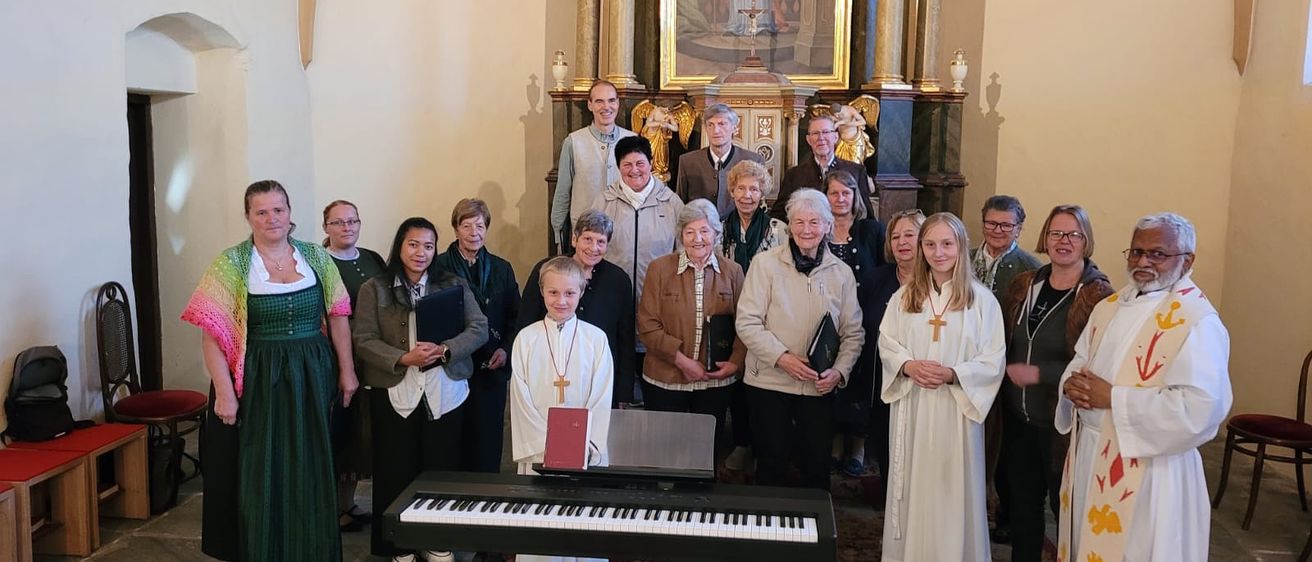 Eine Gruppe von Menschen in verschiedenen Kleidungen, einige mit Büchern, steht vor einem Klavier in einer Kirche, mit einem Gemälde im Hintergrund.