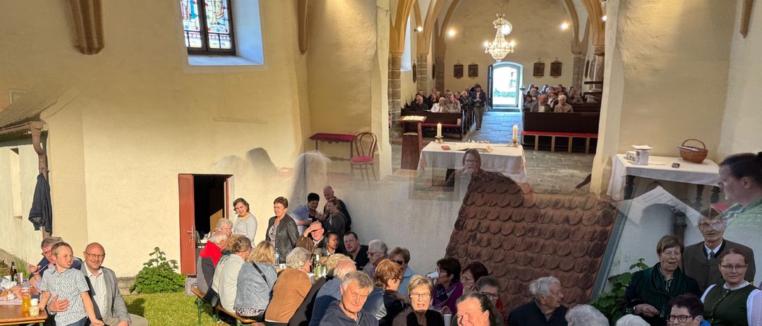 Eine Gruppe von Menschen versammelt sich in einem Kirchensaal, einige sitzen an Tischen, andere stehen. Tische sind mit Bechern, Flaschen und Essen gedeckt. Eine Person in einer grünen und weißen Kleidung steht vor einem Tisch.