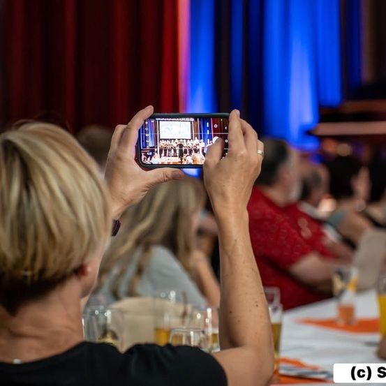 Eine Frau hält ein Smartphone und erfasst eine Aufführung auf der Bühne. Vor ihr sitzen mehrere Menschen und die Bühne ist mit roten und blauen Vorhängen geschmückt.