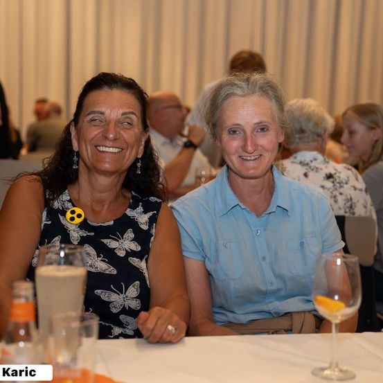 Zwei Frauen lächeln und sitzen an einem Tisch in einem Restaurant. Eine trägt ein Blumenkleid und die andere ein blaues Hemd. Sie sind von anderen Menschen umgeben.