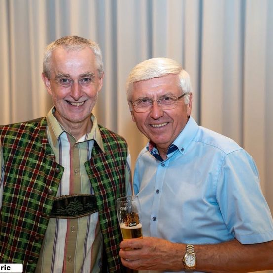 Zwei Männer lächeln und stehen eng beieinander. Einer trägt eine grüne Weste und der andere ein blaues Hemd. Sie halten beide ein Glas Bier.