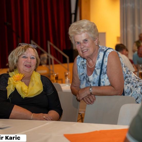 Zwei Frauen sitzen an einem Tisch in einem Speisesaal. Die Frau auf der linken Seite trägt ein gelbes Tuch mit einer Blume. Die Frau auf der rechten Seite hat eine Handtasche und einen Armreif. Hinter ihnen ist ein roter Vorhang.