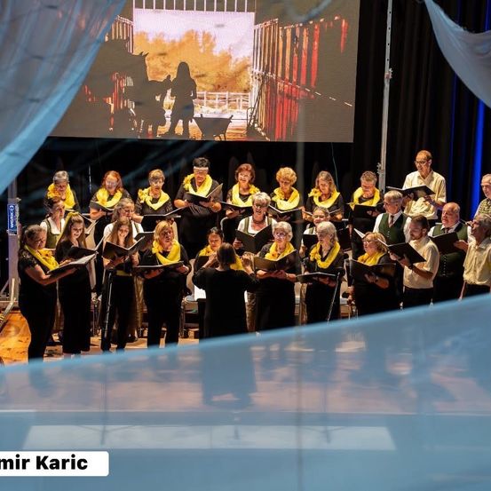 Ein Chor tritt auf der Bühne auf, während eine große Leinwand eine ländliche Szene im Hintergrund zeigt.