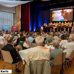 Ein Chor tritt auf der Bühne auf, im Hintergrund ein großer Bildschirm. Zuschauer sitzen an Tischen, klatschen und lächeln.