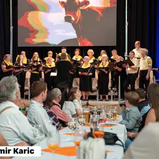 Ein Chor tritt auf der Bühne auf, mit einer Kuh, die auf einer Leinwand hinter ihnen projiziert wird. Menschen sitzen davor und beobachten und trinken Bier.