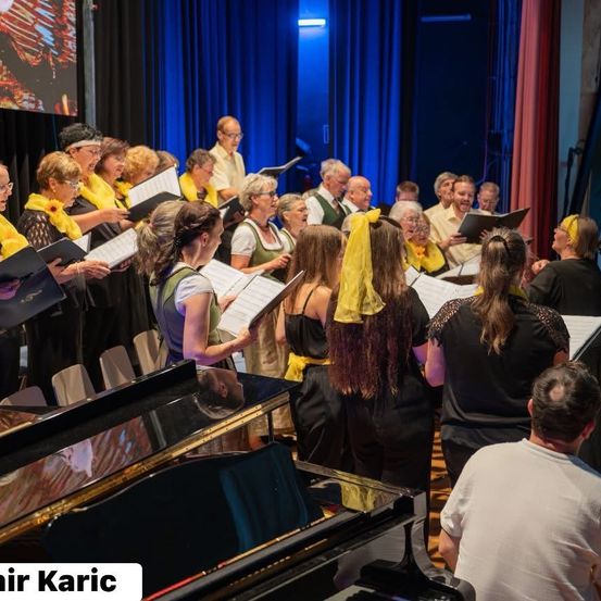 Ein Chor tritt auf der Bühne mit blauen Vorhängen und einer Leinwand auf. Das Publikum sitzt im Hintergrund. Die Gruppe von Menschen hält Notenblätter und singt.