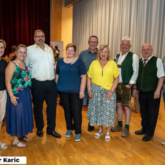 Eine Gruppe von Menschen in verschiedenen Outfits, möglicherweise für ein besonderes Ereignis, posiert für ein Foto in einem Raum mit Holzböden und einem roten Vorhang.