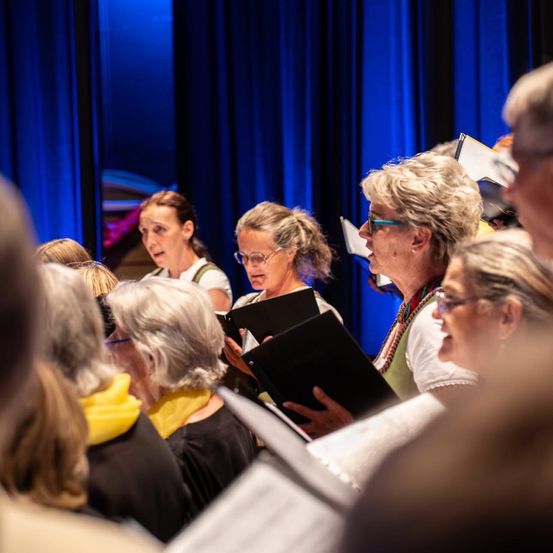 Eine Gruppe älterer Frauen singt gemeinsam und hält Notenblätter in den Händen. Sie stehen auf einer Bühne mit blauen Vorhängen im Hintergrund.