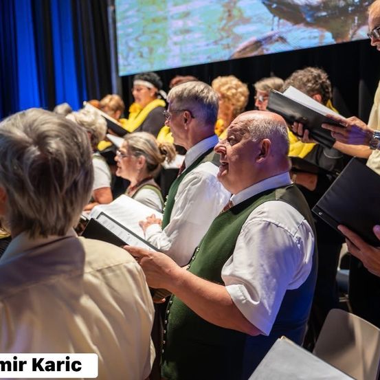 Eine Gruppe von Menschen singt in einem Chor, hält ihre Notenblätter in den Händen und steht vor einer großen Leinwand.