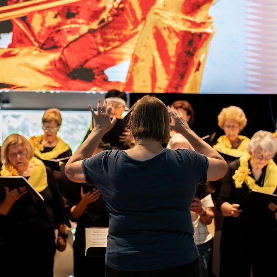 Ein Chor von Frauen, geschmückt mit gelben Schärpen, singt in einem großen Raum mit einer goldenen Figur auf einem Bildschirm.