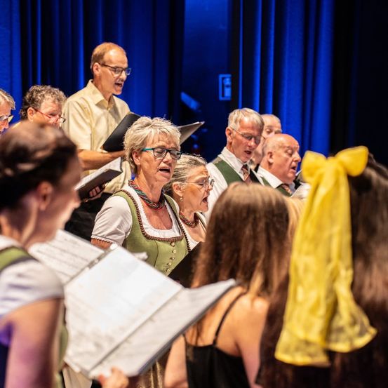 Ein Chor älterer Erwachsener in traditioneller Kleidung singt auf der Bühne vor blauen Vorhängen. Einige halten offene Bücher, während andere zusehen.