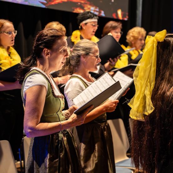 Eine Gruppe von Frauen in traditionellen Kostümen singt in einem Chor und hält Notenblätter. Eine Frau sticht mit einem gelben Band hervor.