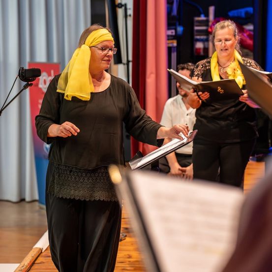 Eine erwachsene Frau mit einem gelben Schal geht vorwärts und hält ein Musikbuch. Eine andere Frau mit einem gelben Schal liest aus einem Buch. Ein Mann sitzt hinter ihnen auf dem Boden.
