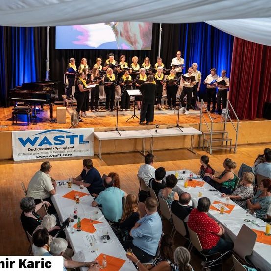 Ein Chor tritt auf einer Bühne auf, mit einem Pianisten und einer Leinwand hinter ihnen. Das Publikum sitzt an Tischen vor der Bühne. Die Bühne hat ein Banner, das Wastl liest.