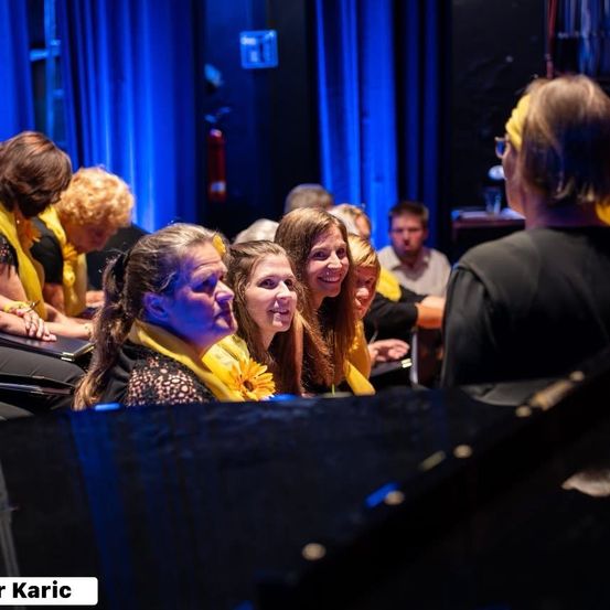 Eine Gruppe von Frauen mit gelben Tüchern mit Sonnenblumen sitzt auf Stühlen in einem Theater, mit einem Klavier im Vordergrund.