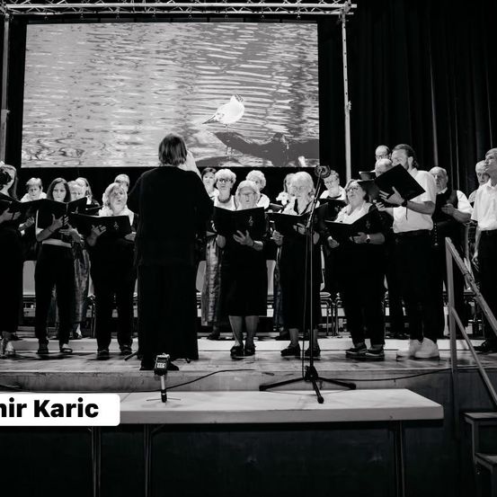 Ein Chor tritt auf der Bühne auf, mit einer großen Leinwand im Hintergrund, die einen Vogel zeigt. Alle Mitglieder halten Bücher und scheinen zu singen.