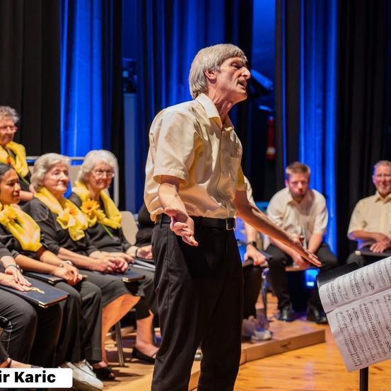 Ein Mann steht auf der Bühne mit einem Notenständer vor ihm. Er singt für einen Chor, der in Stühlen sitzt.