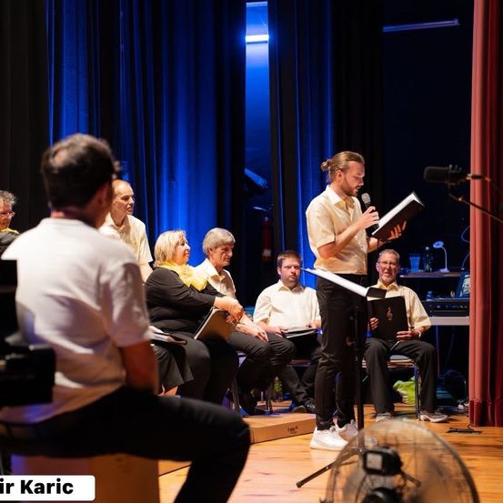 Ein Chorprobe mit einem Mann, der in ein Mikrofon spricht, während andere aufmerksam zuhören. Die Umgebung umfasst eine Bühne mit blauen Vorhängen und einem Holzboden.
