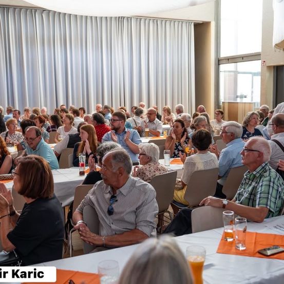Eine Gruppe von Menschen sitzt in einem Raum an Tischen und klatscht. Einige Leute tragen Brillen und andere Armbänder. Ein Tisch hat Gläser, eine Flasche und ein Handy. Ein Wasserzeichen befindet sich unten links.