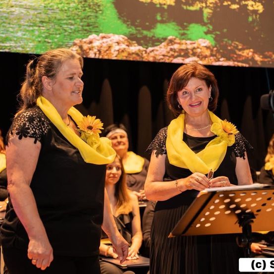 Zwei Frauen in schwarzen Kleidern und gelben Schals mit Blumen stehen vor einem Podium, wahrscheinlich bei einer Zeremonie. Ein Publikum sitzt hinter ihnen. Eine große Leinwand zeigt eine landschaftliche Kulisse.