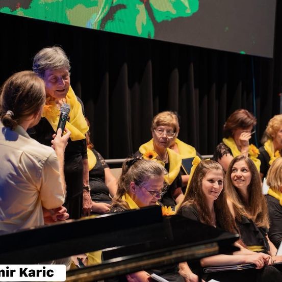 Ein Chor von Frauen in gelben Schals singt in einem Konzertsaal, eine Frau hält ein Mikrofon und der Bildschirm hinter ihnen zeigt ein grünes Blatt.