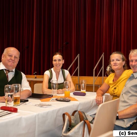 Fünf Personen, drei Männer und zwei Frauen, sitzen an einem Tisch mit Gläsern und Essen und lächeln in die Kamera. Hinter ihnen sind rote Vorhänge und eine Treppe zu sehen.