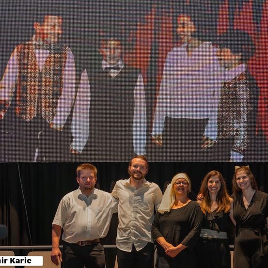 Fünf Personen stehen vor einer großen Leinwand und lächeln für ein Foto. Dahinter wird eine Gruppe von Männern auf dem Bildschirm gezeigt, die formelle Kleidung tragen.