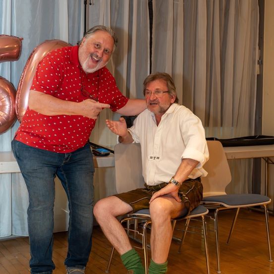 Zwei Männer sind in einem Raum. Ein Mann steht und zeigt, der andere sitzt auf einem Stuhl. Ein Luftballon mit der Nummer 50 steht hinter ihnen.